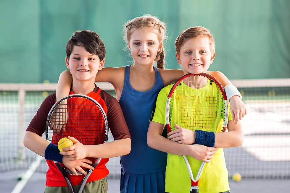 3 fröhliche Kinder mit Tennisschlägern in der Hand lächeln in die Kamera.