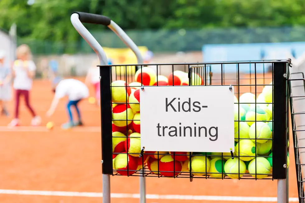 Ballwagen gefüllt mit bunten Tennisbällen und einem Schild mit der Aufschrift: Kids-Training. Kinder spielen im Hintergrund Tennis.
