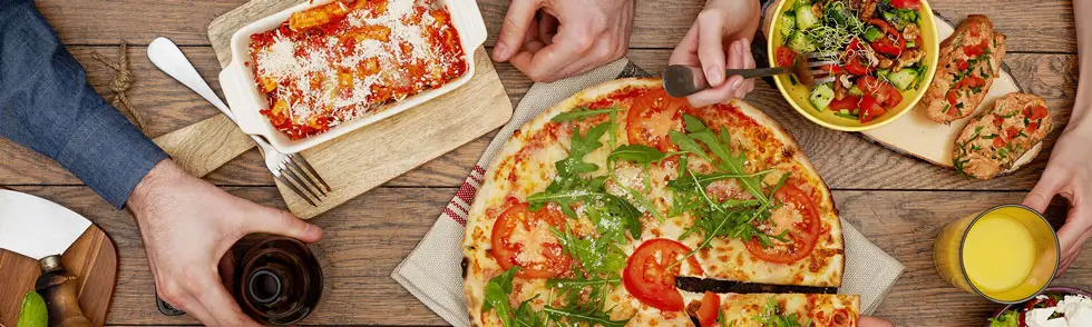 mehre Personen beim Essen. Auf dem Tisch stehen italienische Spezialitäten: Lasagne, Pizza, Salate, Bruschetta
